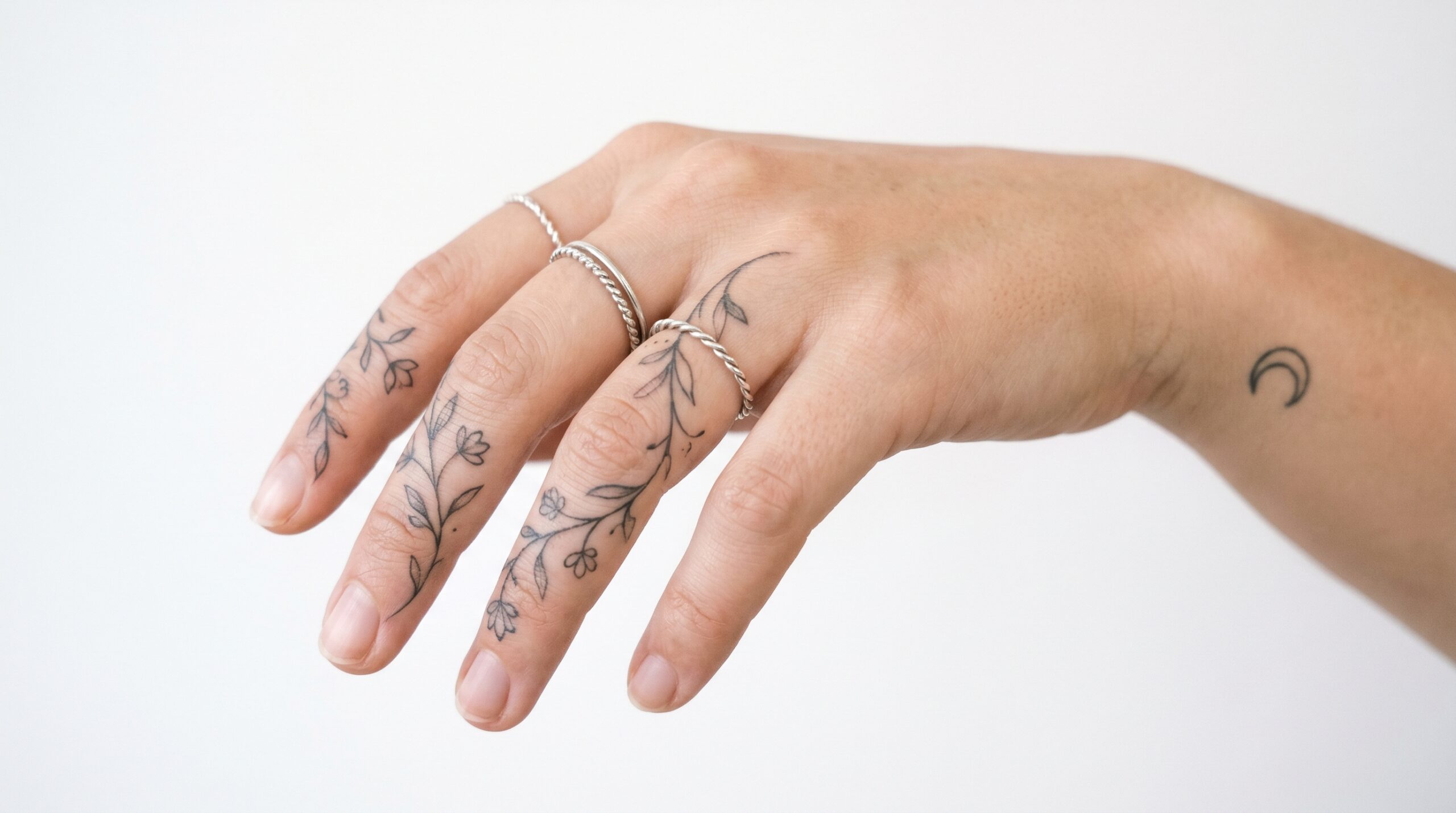 *Primo piano elegante della mano di una donna con delicati tatuaggi fine-line, motivi ornamentali a forma di vite lungo indice e medio e una piccola luna crescente sul lato del polso.*
