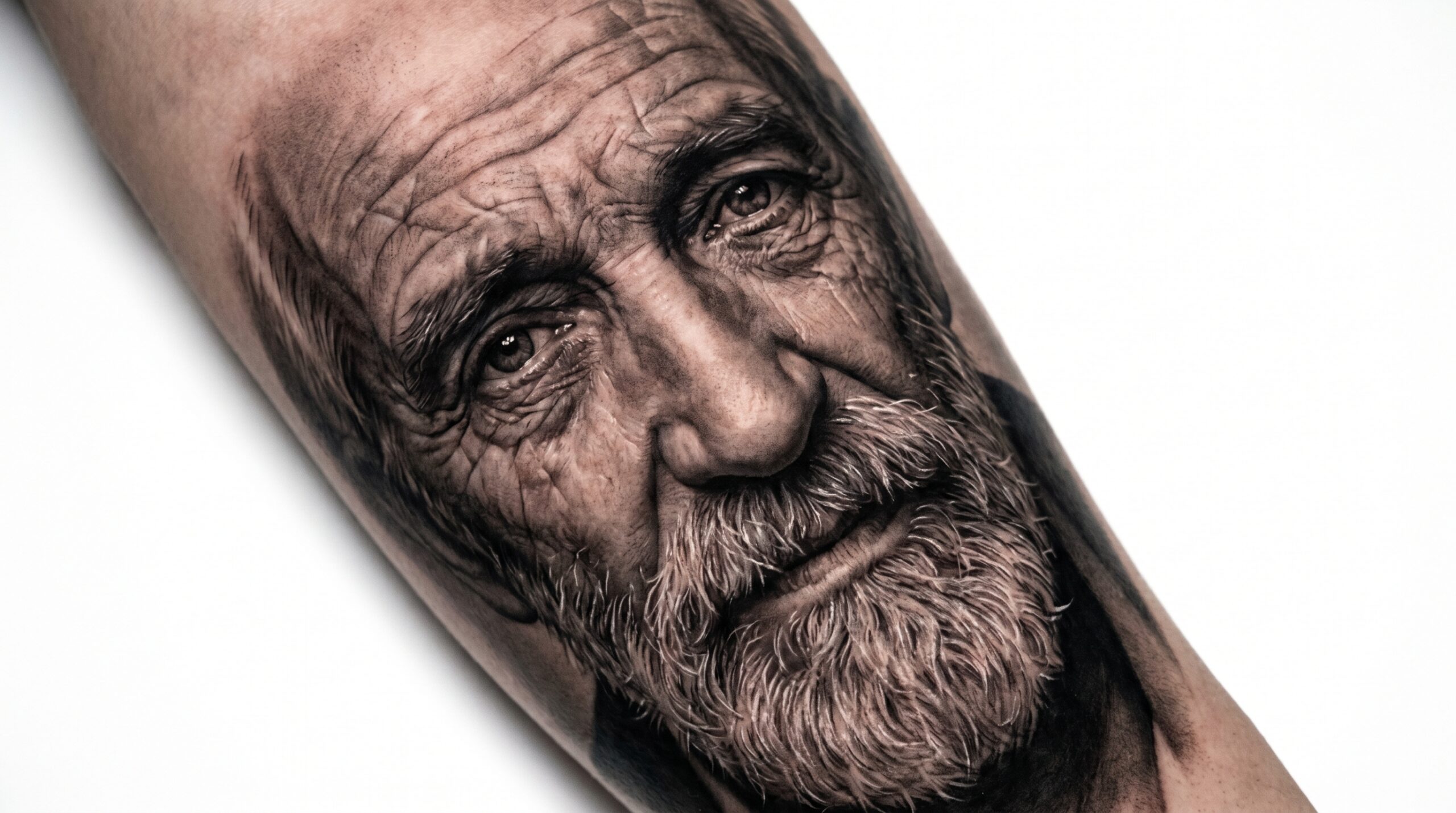 "Tatuaggio ritratto iperrealistico Black and Grey su braccio raffigurante un uomo anziano con barba grigia dettagliata, sguardo realistico, rughe delicate e sfumature morbide senza contorni marcati, in fotografia macro su sfondo bianco."