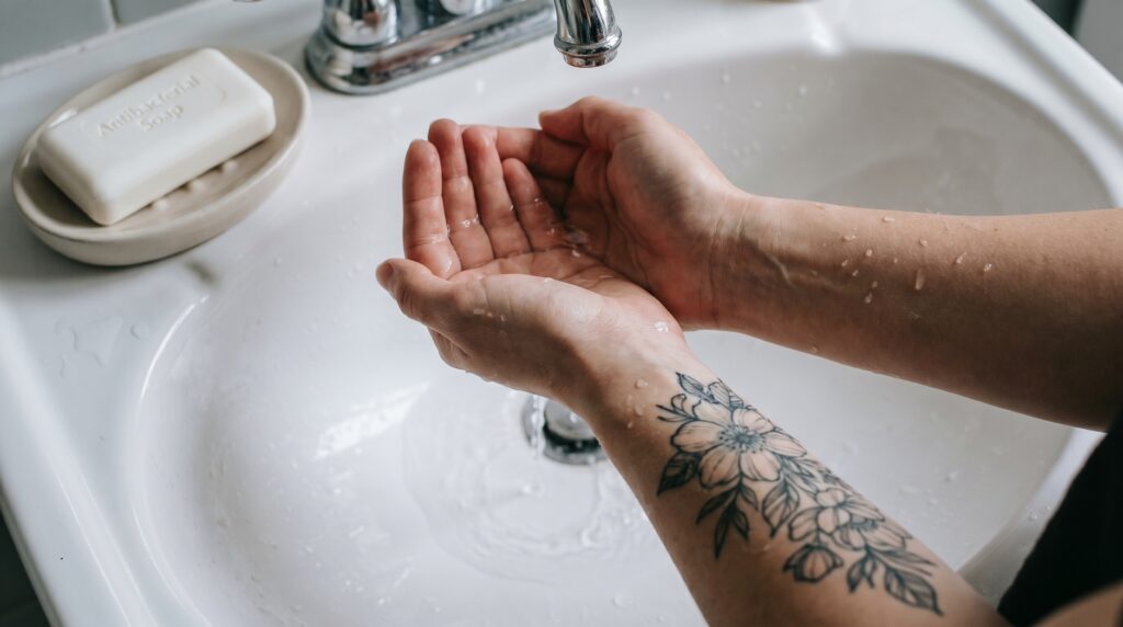 Una ripresa in prima persona sopra un lavandino del bagno. Delle mani tengono a coppa dell'acqua tiepida per sciacquare delicatamente un tatuaggio appena fatto. Una saponetta neutra e antibatterica è visibile di lato. L'immagine dovrebbe trasmettere un senso di delicatezza, pulizia e cura.