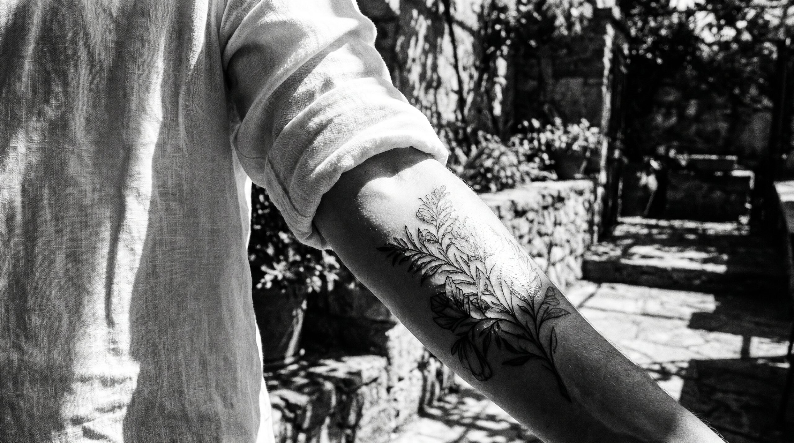 Fotografia ad alto contrasto del braccio di una persona con un tatuaggio appena fatto, parzialmente ombreggiato da una leggera camicia di lino. Metà del tatuaggio è nascosta dal tessuto, mentre l'altra metà è colpita da un raggio di sole forte e abbagliante, a rappresentare visivamente la fragilità dell'inchiostro. Sullo sfondo si vede una luminosa terrazza estiva.