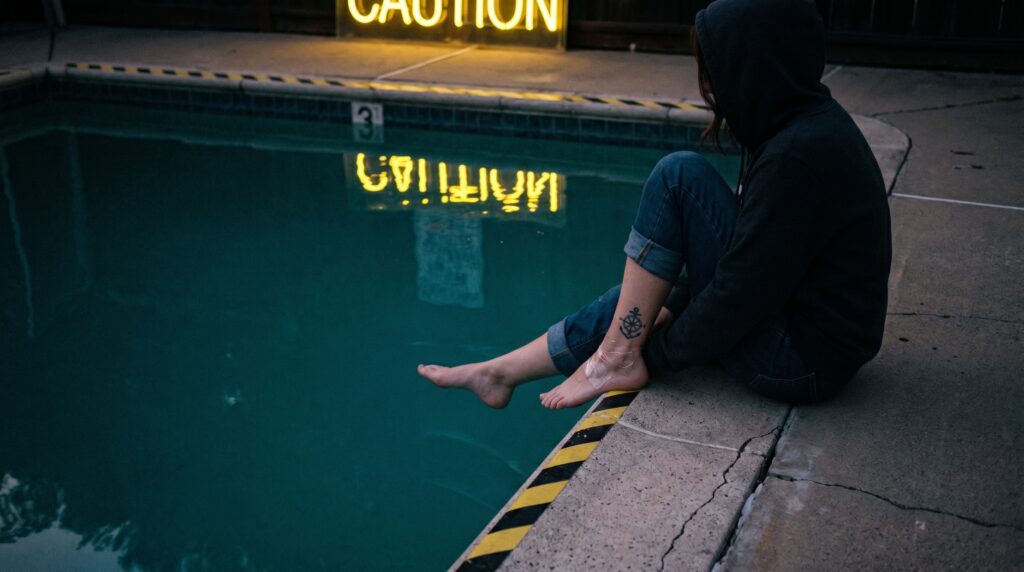 Un'immagine suggestiva e malinconica di una persona seduta sul bordo di una piscina turchese, con i piedi che penzolano appena sopra l'acqua. Un'attenzione particolare è rivolta a un tatuaggio fresco sulla caviglia, asciutta e al sicuro dall'acqua clorata. L'acqua stessa evoca un senso di "prudenza".