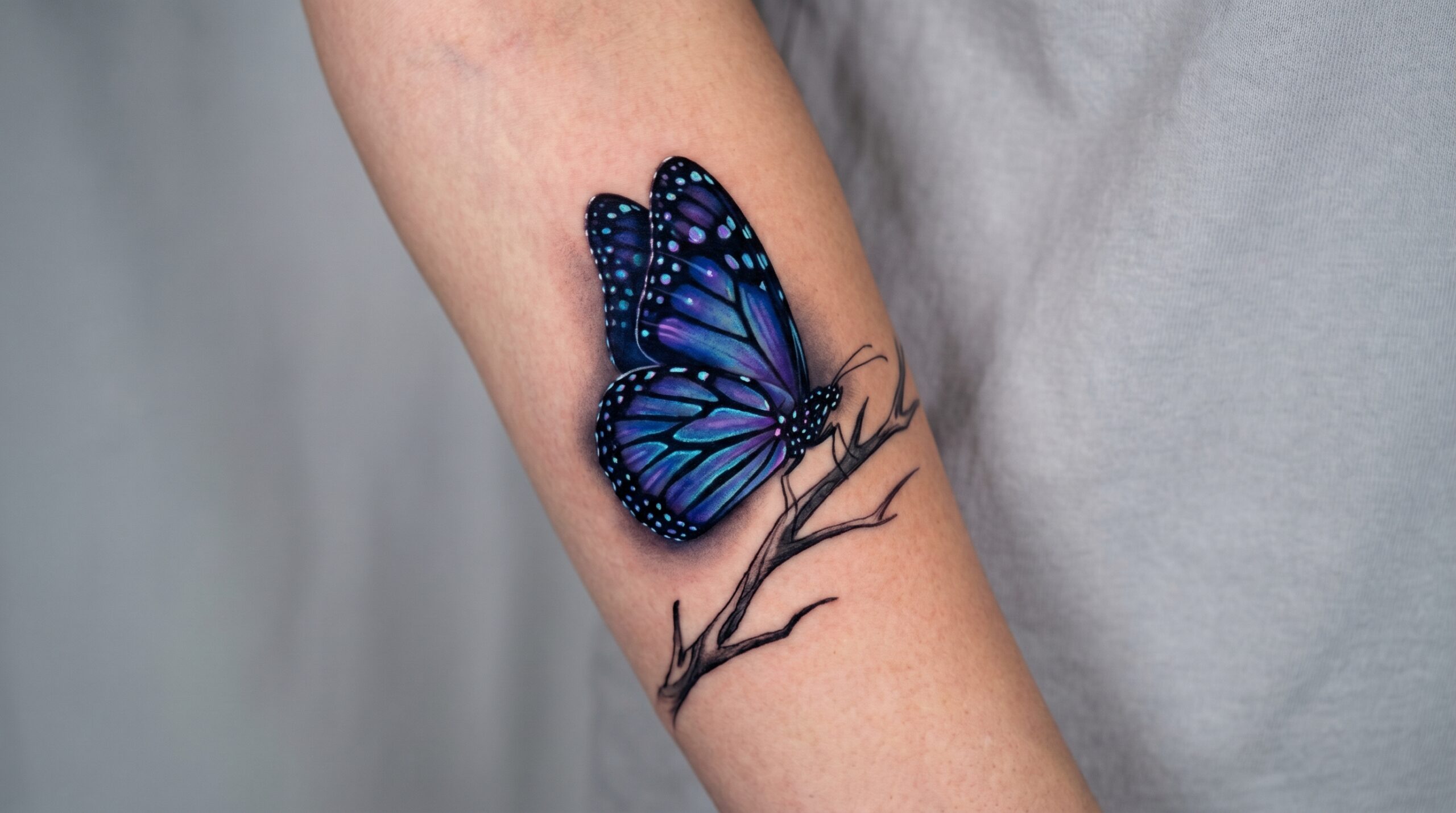 Tatuaggio realistico raffigurante una farfalla monarca con ali di un vibrante blu cobalto e indaco al posto del classico arancione. La texture dettagliata delle ali, i micro-brillanti riflessi luminosi e le delicate sfumature creano un effetto tridimensionale, come se la farfalla fosse posata su una superficie.