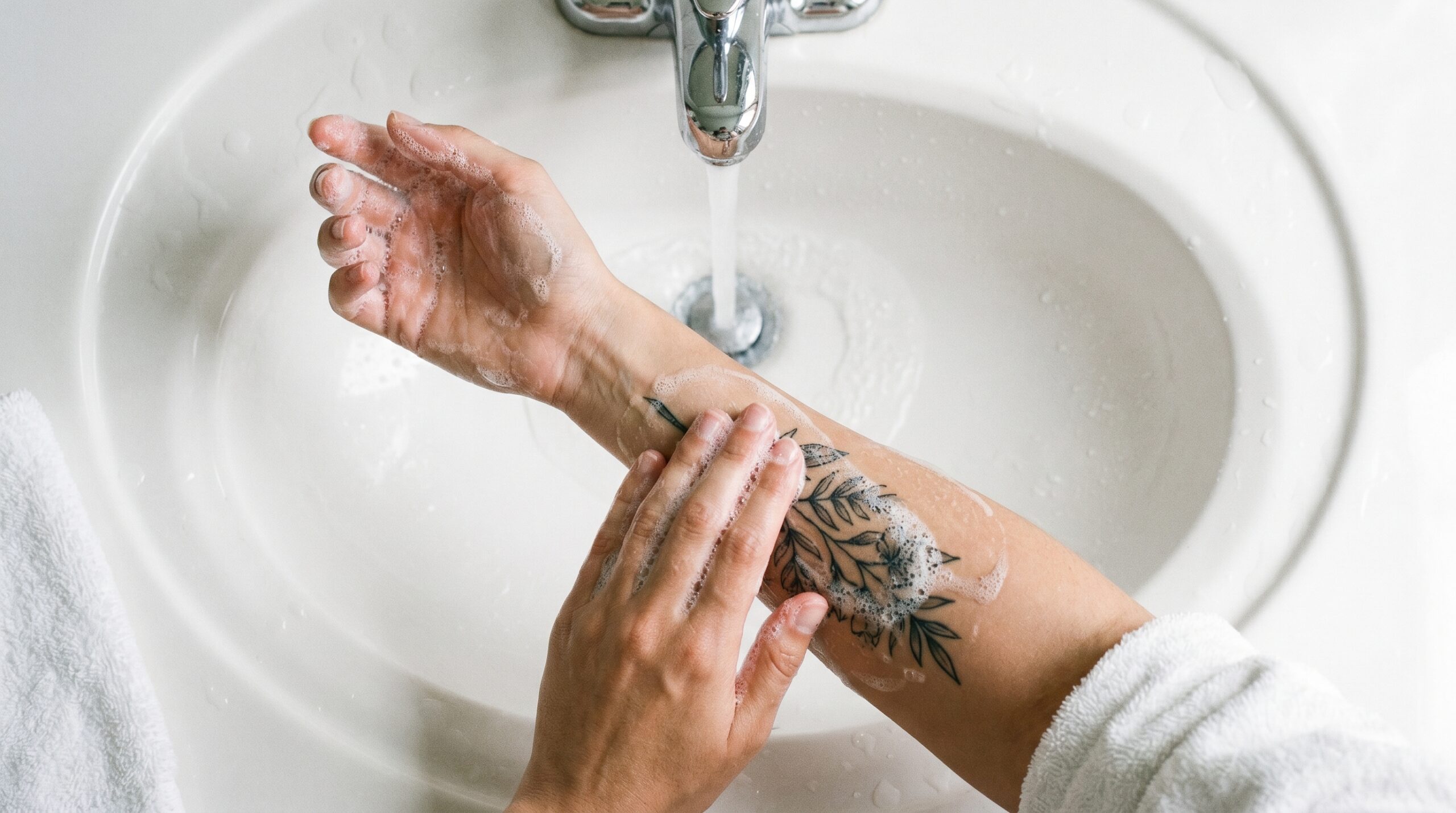 Una persona sta lavando delicatamente un nuovo tatuaggio sull'avambraccio nel lavandino del bagno. Utilizza la punta delle dita con movimenti circolari e un sapone delicato, bianco e schiumoso. L'acqua tiepida scorre dolcemente.